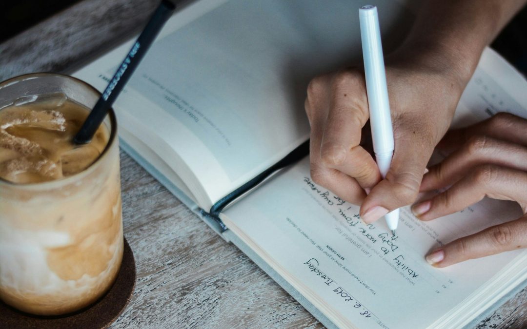 Hand schreibt in ein aufgeschlagenes Notizbuch, daneben ein Glas Eiskaffee