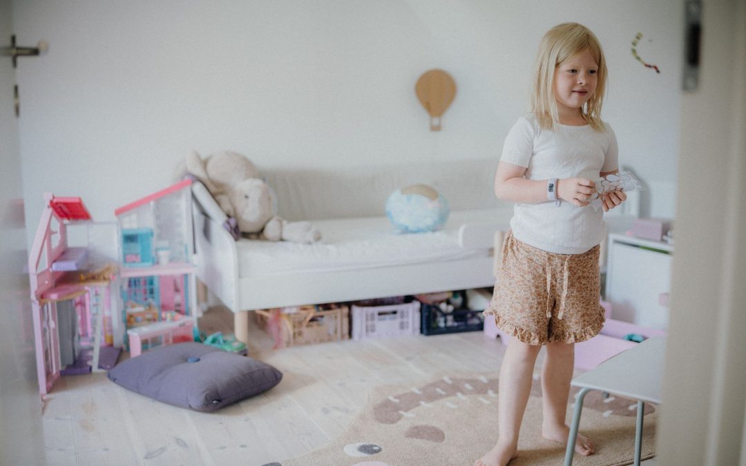 Mädchen steht in ihrem Kinderzimmer, im Hintergrund Bett mit Stauraumkisten, Puppenhaus und Spielteppich