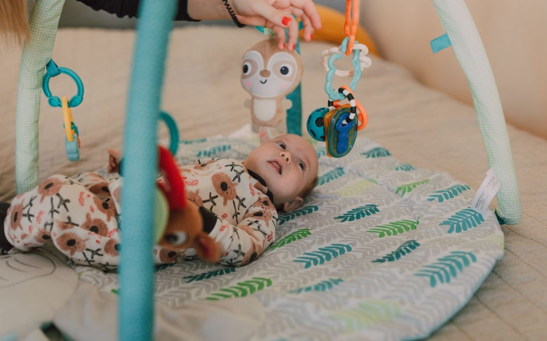 Baby liegt auf einer gepolsterten Krabbeldecke unter einem Spielbogen mit Anhängern