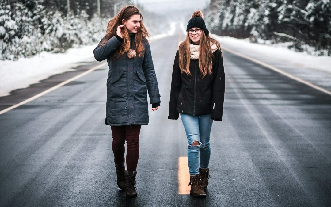 Zwei junge Frauen gehen nebeneinander über eine verschneite Straße zwischen Tannen