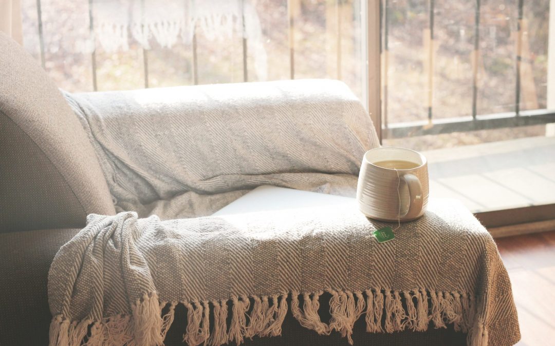 Sofa mit dicker Decke und einer Tasse Tee, dahinter ein Fenster mit kahlen Bäumen