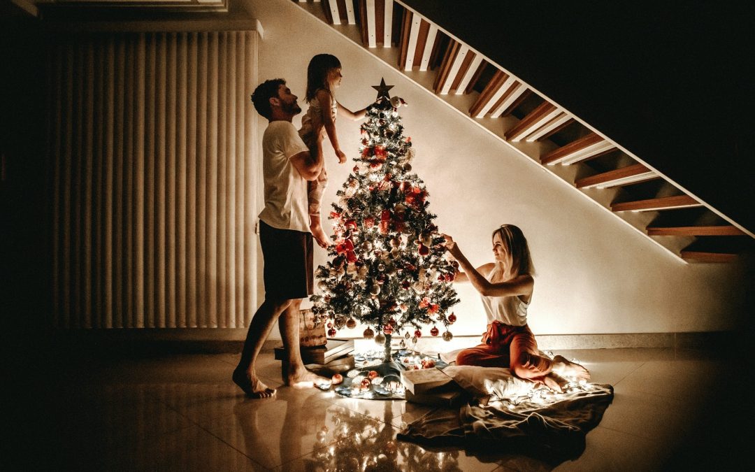 Vater hebt ein Kind hoch, das den Stern auf den Weihnachtsbaum setzt, die Mutter hängt Kugeln auf
