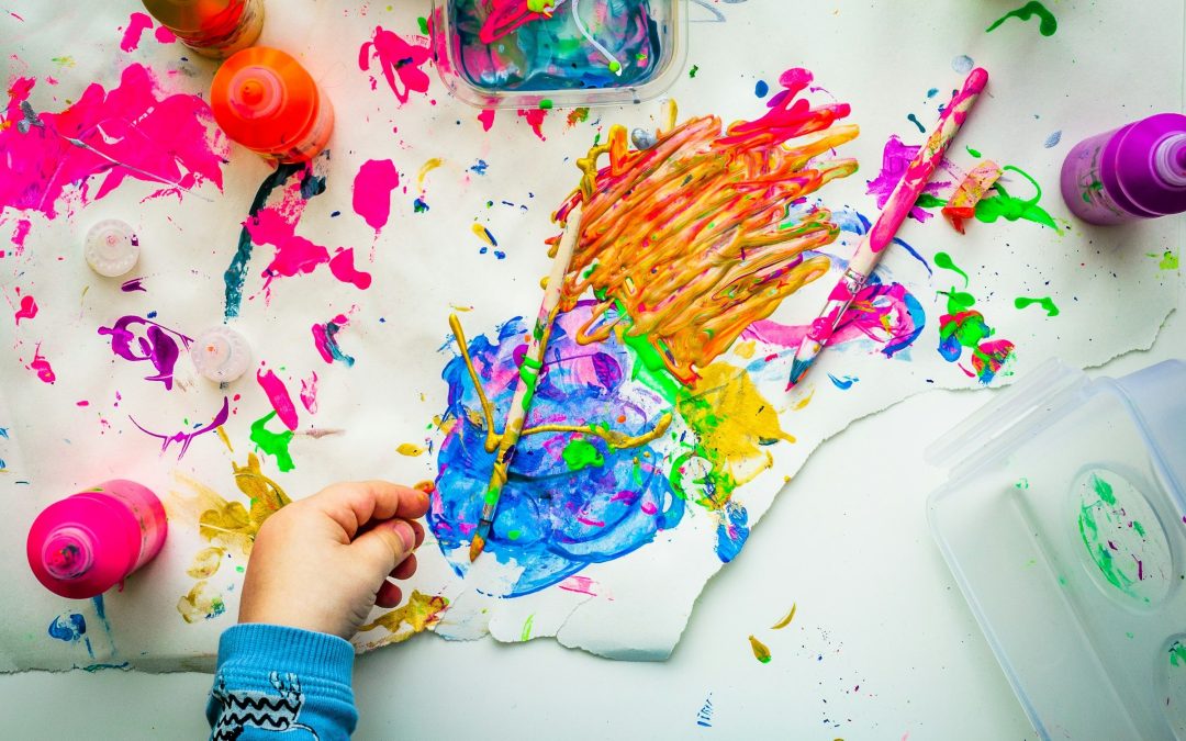 Eine Kinderhand im blauen Aermel haelt einen Pinsel ueber ein Blatt voller bunter Fingerfarbe, daneben Farbflaschen